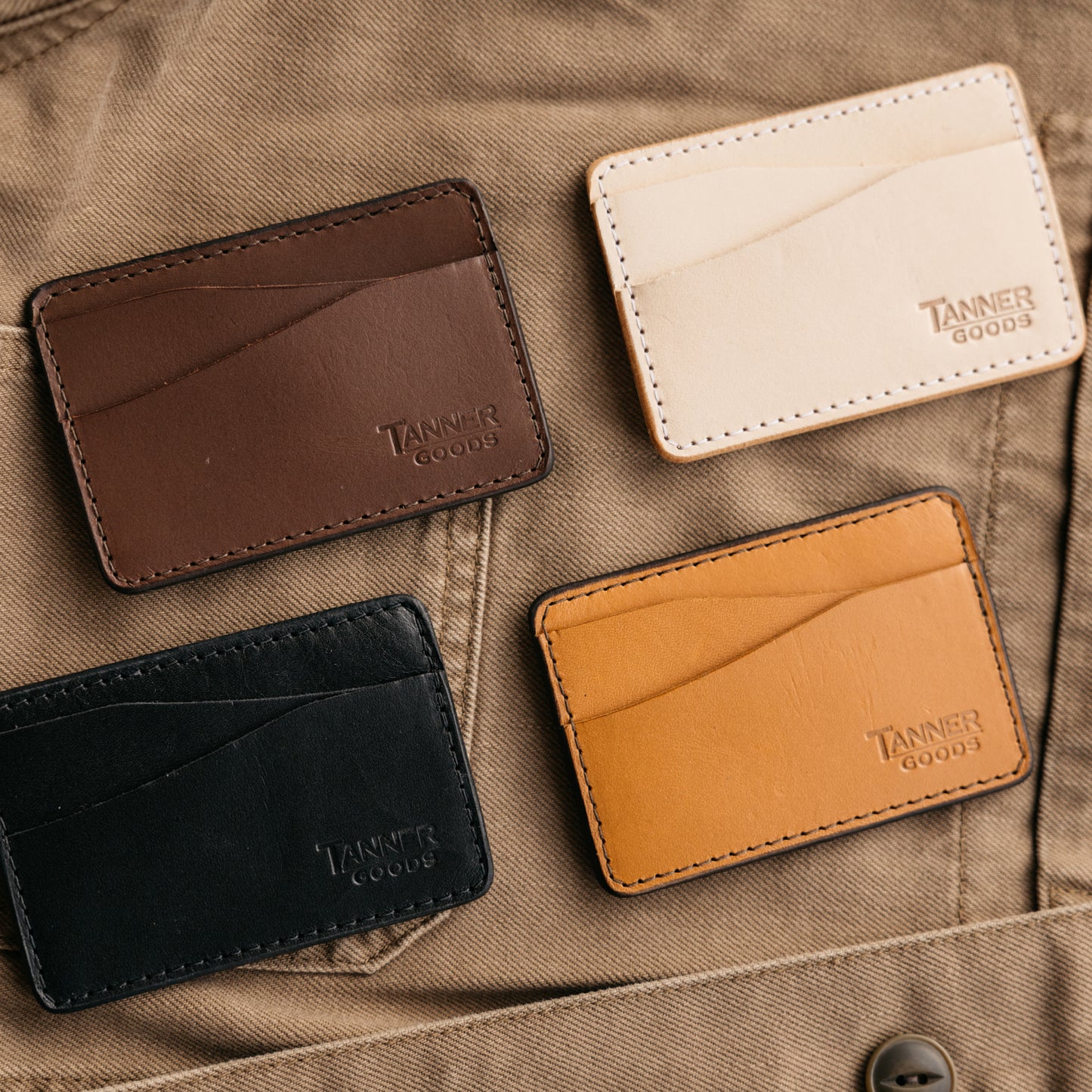 Black leather Money Clip Journeyman wallet by Tanner Goods, featuring embossed logo and visible stitching, displayed with three similar wallets on khaki fabric.