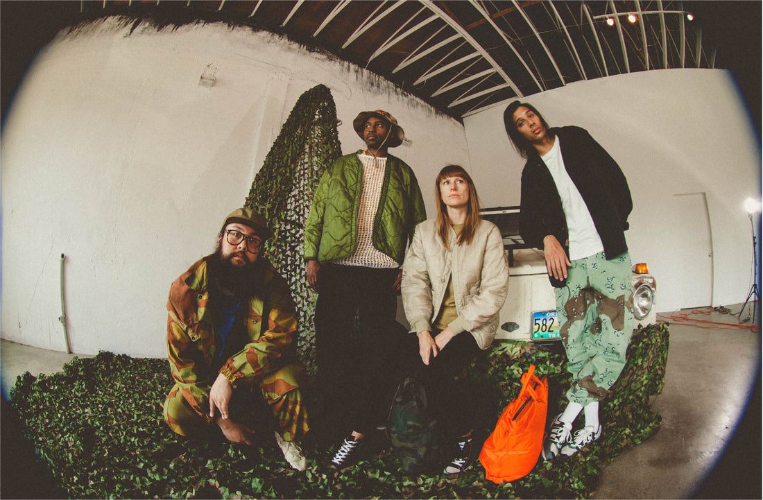 Four people in eclectic, nature-inspired outfits pose in a warehouse with camouflage netting and a vintage truck, evoking a raw, artistic vibe.