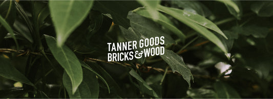 Lush green foliage backdrop with bold white text "TANNER GOODS BRICKS&WOOD" centered, evoking natural craftsmanship and collaboration.