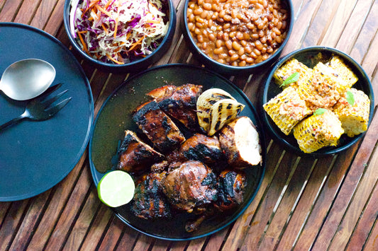 Grilled chicken, corn, beans, and coleslaw on dark plates, served on a wooden table with lime and garlic.