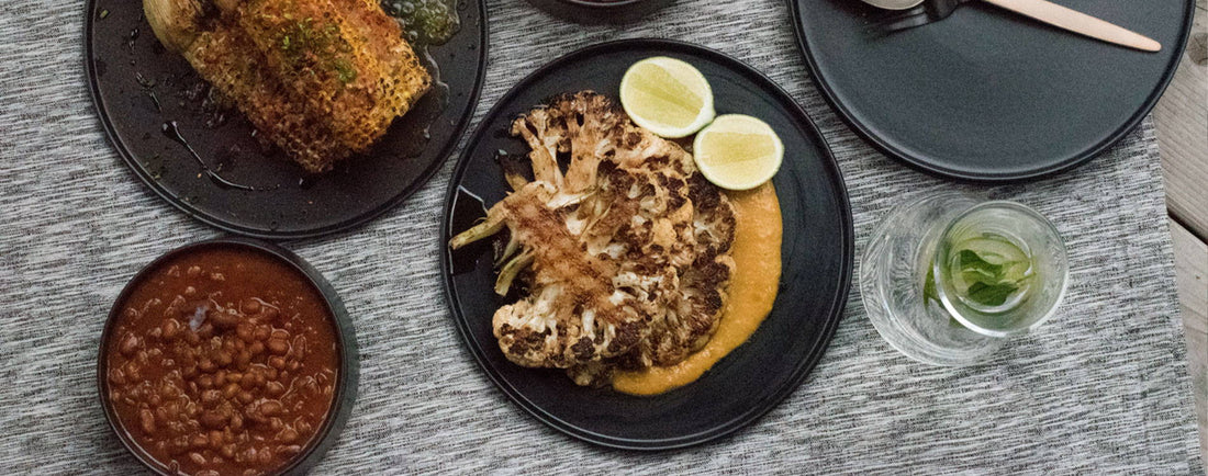 Roasted cauliflower steak with lime, beans, and crispy fish on dark plates, set on a textured gray tablecloth.