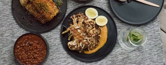 Roasted cauliflower steak with lime, beans, and crispy fish on dark plates, set on a textured gray tablecloth.