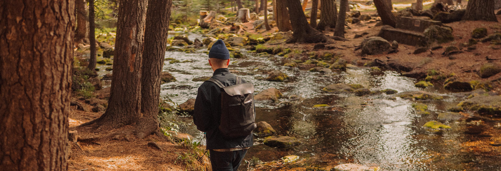 Man in forest with rugged backpack and beanie, exploring nature trail near stream — The Explorer collection.