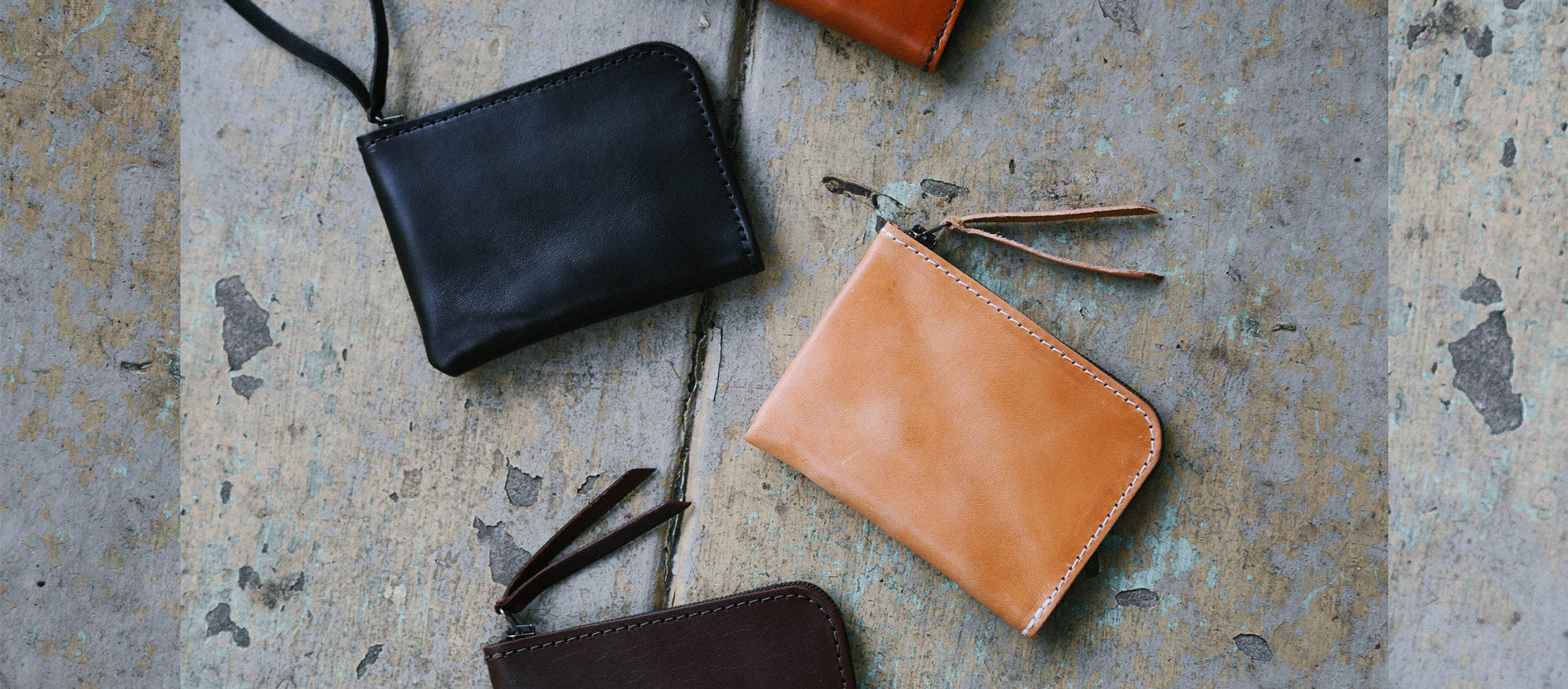 Minimal leather zip wallets in black, tan, and brown with contrast stitching on weathered concrete.