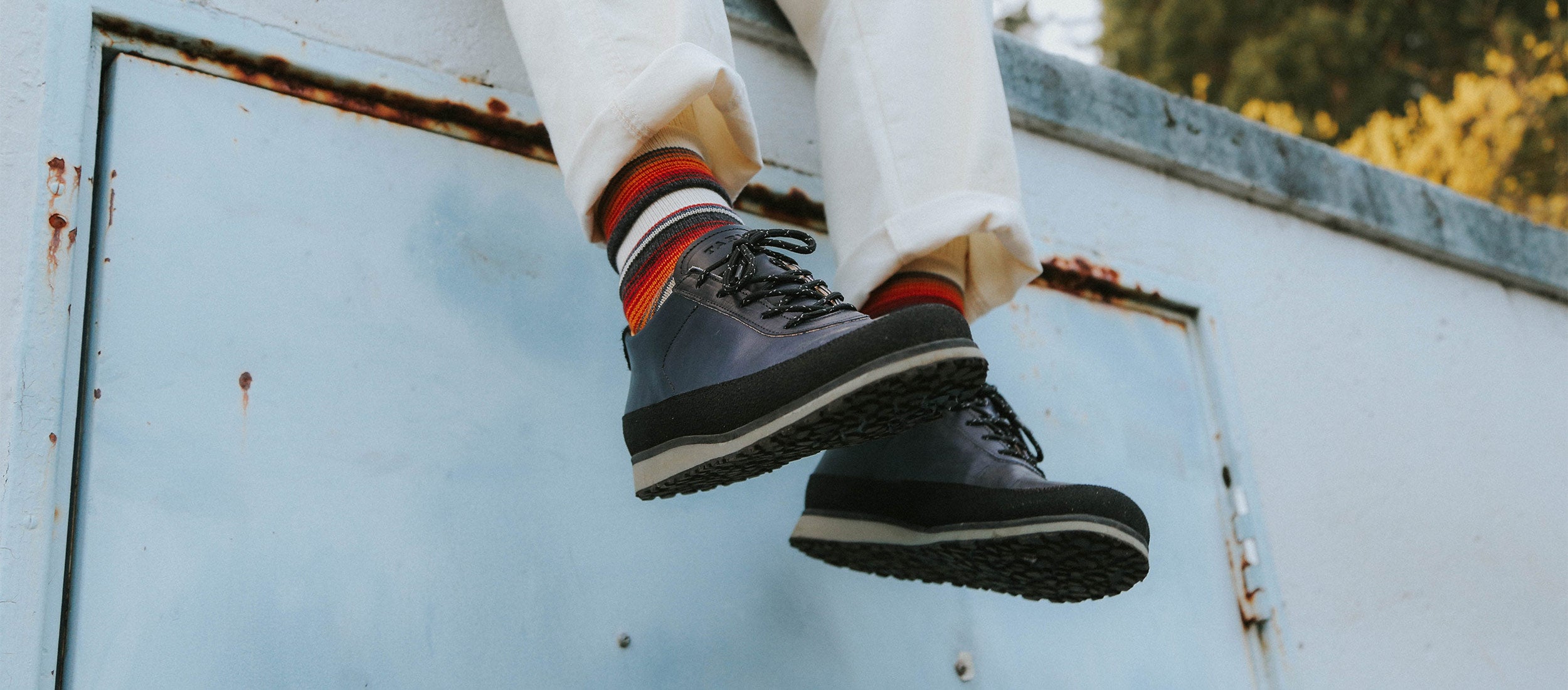 Stylish navy and black lace-up shoes with bold striped socks, white cuffed pants, against a weathered blue door—urban, relaxed, confident footwear.