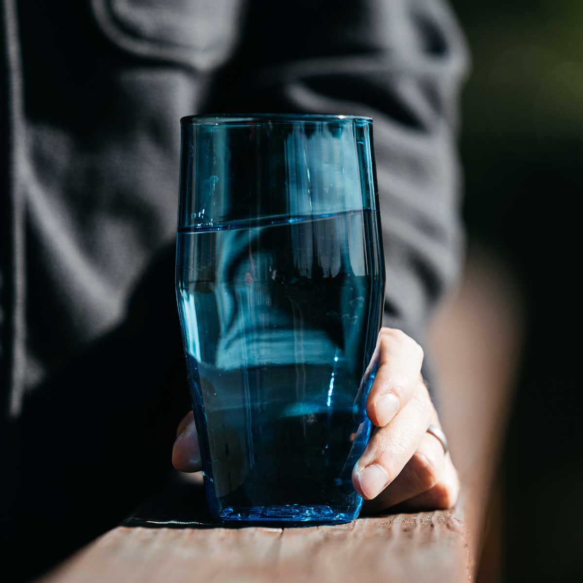 Blue Standard Pint Glass filled with water, held by a hand resting on a wooden railing.