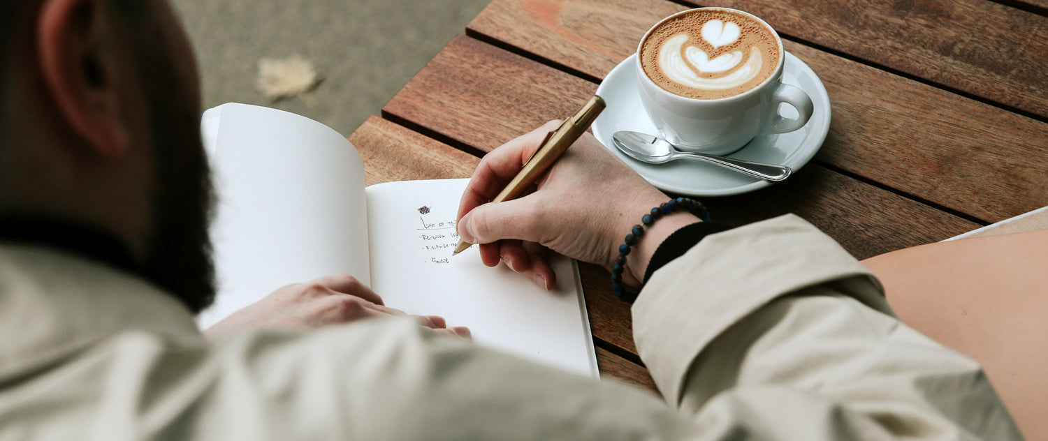 Man writing in notebook at wooden table with latte art coffee, golden pen, and beaded bracelet.