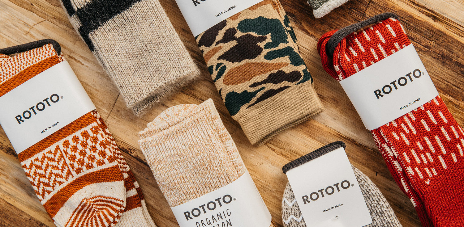 Colorful ROTOTO socks with patterns like camouflage and stripes, labeled "Made in Japan," arranged on a wooden surface.