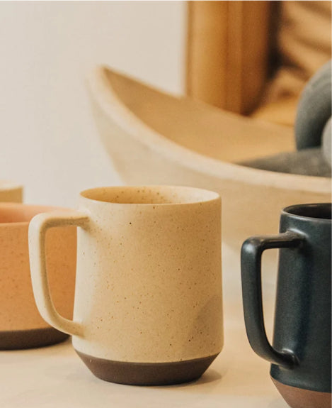 Earthy-toned ceramic mugs with speckled beige and dark green glazes, arranged on a light surface with blurred wooden bowls in the background.