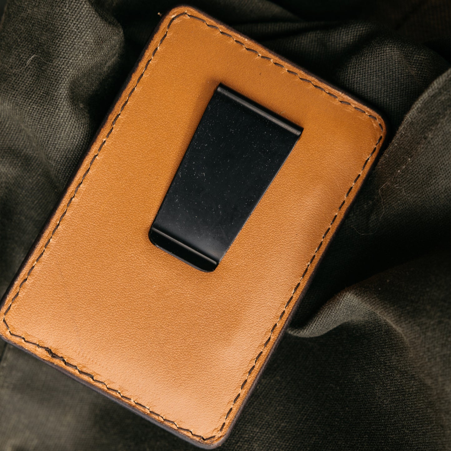 Black money clip and tan leather card holder with stitched edges, lying on dark green textured fabric.