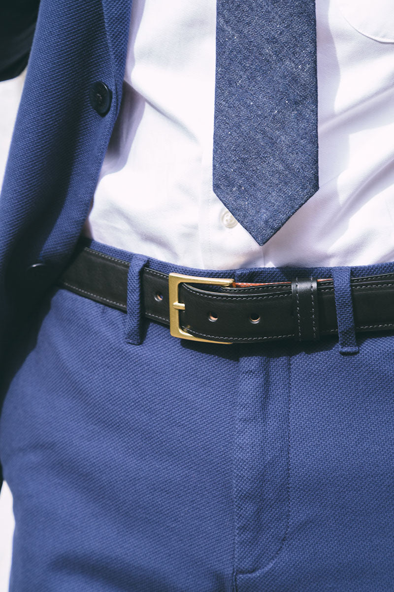 Black leather dress belt with a gold buckle, worn with blue textured trousers, a white shirt, and a blue blazer.